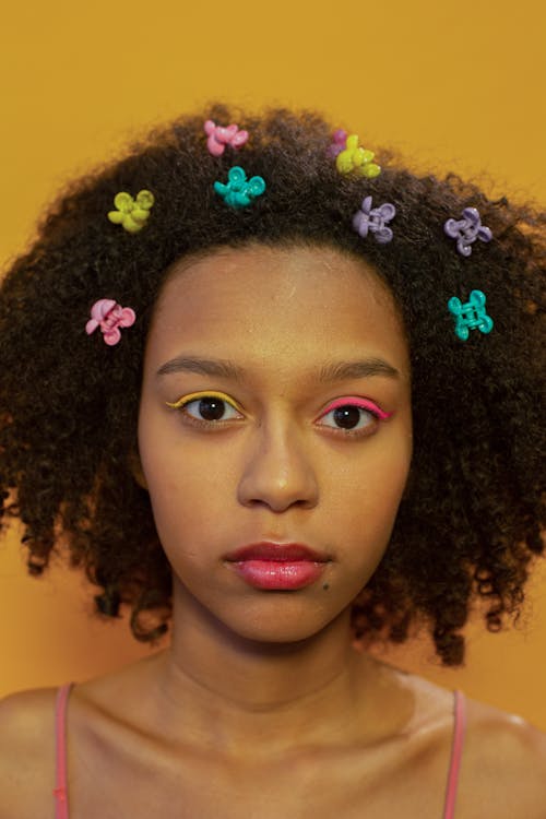 Emotionless African American teenager with colorful eyeshadows and small hairpins looking at camera on yellow background in light studio