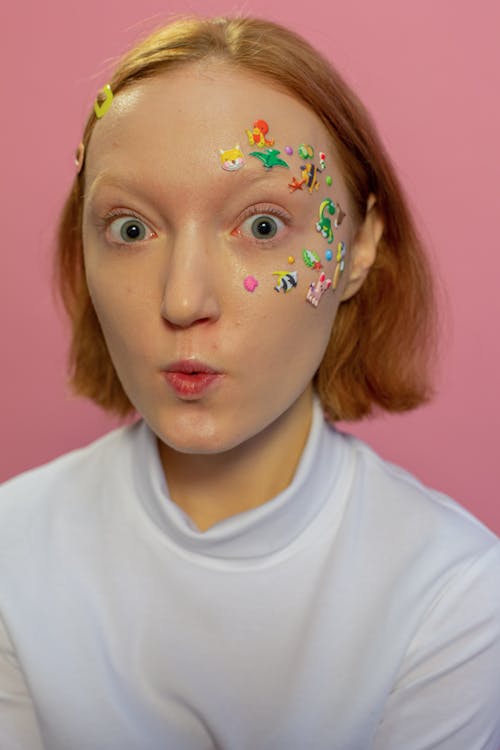 Amazed young lady in casual clothes and colorful animal stickers on face with hairpins in hair looking at camera on pink background in light studio