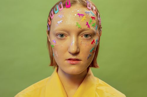 Free Serious young lady with colorful unicorn stickers and glitter on face with hairpins in hair looking at camera on green background in light studio Stock Photo