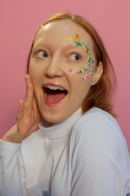 Shocked young woman in casual outfit and colorful stickers in shape of animals on face and hairpins in hair looking away with open mouth in bight studio on pink background