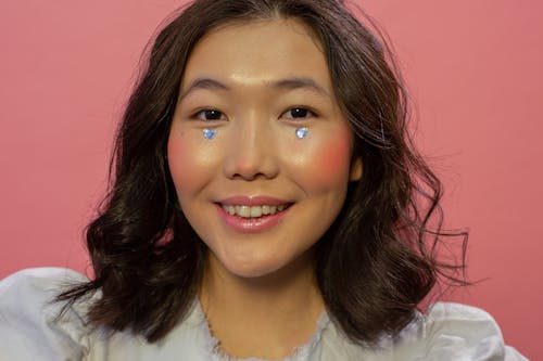 Happy young Asian woman with shimmering rhinestones in shape of hearts on cheeks and makeup looking at camera in bight studio on pink background