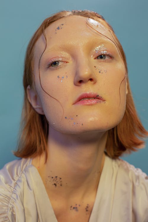 Emotionless young female model with ginger hair and glossy glitter on face looking away against blue background in studio