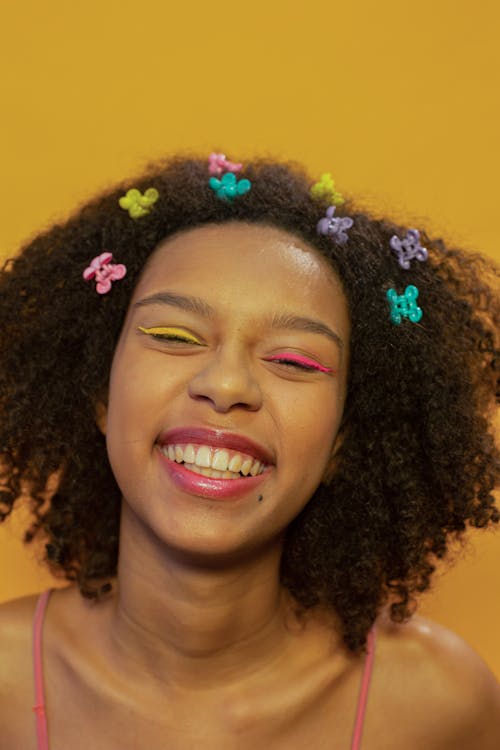 Delighted ethnic female teenager with colorful makeup and mini claws in dark Afro hair smiling happily against yellow background