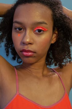 Confident young black female model with bright creative makeup touching curly Afro hair and looking at camera against blue background