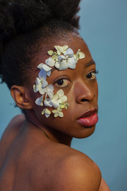 Free Side view gorgeous African American female with bare shoulders and tender blue flower petals on face standing on blue background and looking at camera Stock Photo