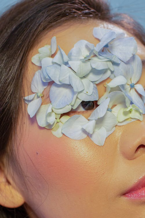 Crop young attractive Asian female with tender blue flower petals on face against blue background