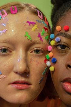 Crop unemotional multiethnic female models with colorful stickers and fluffy balls on face looking at camera against green background