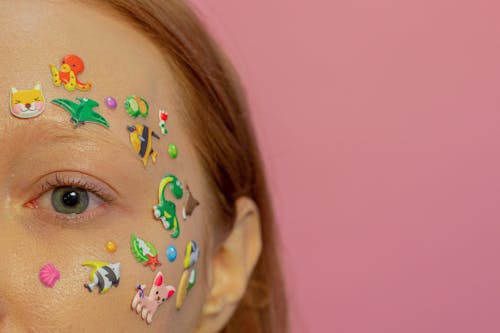 Half face of crop redhead female with blue eyes and multicolored stickers on face against pink background