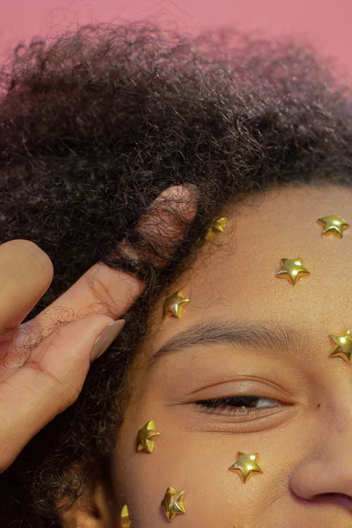 Crop happy African American female with golden stars on face skin touching curly hair and looking at camera in studio