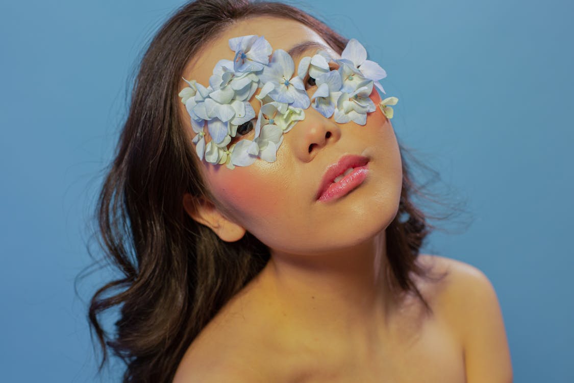 Young attractive female with bare shoulders and tender flower petals on face looking at camera against blue background in studio