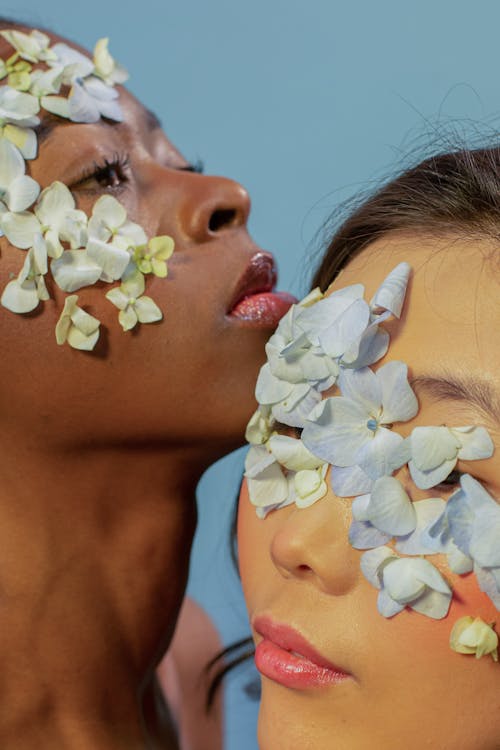 Crop young Asian and African American female models with fresh flower petals on faces against blue background