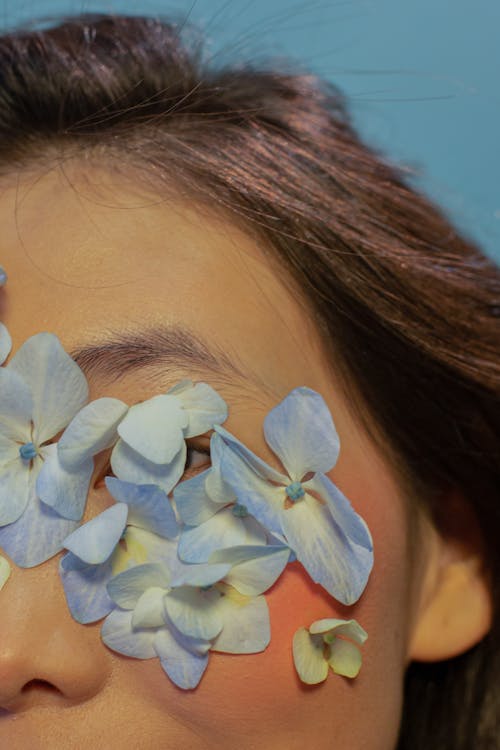 Crop anonymous woman with delicate flower petals on eyes in blue studio