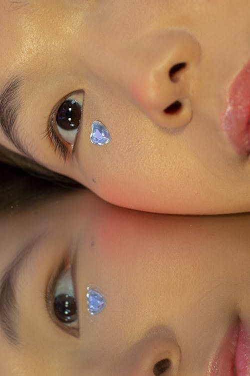 Crop young ethnic lady with perfect skin and shiny rhinestone on face lying on mirror and looking away