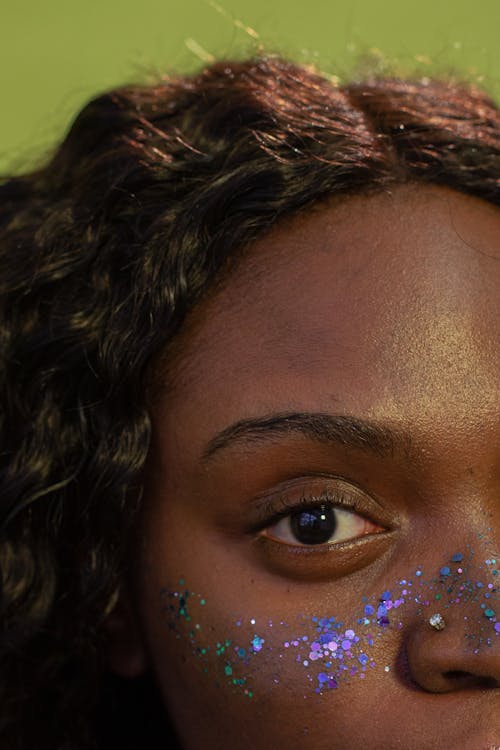 Crop unemotional African American female with wavy hair shiny blue glitters on pretty face looking at camera against green background