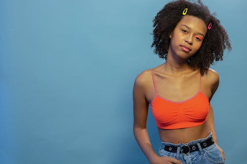 Young glad African American female model with vivid orange eyeshadows on one eye wearing casual wear standing on blue background in studio and looking at camera