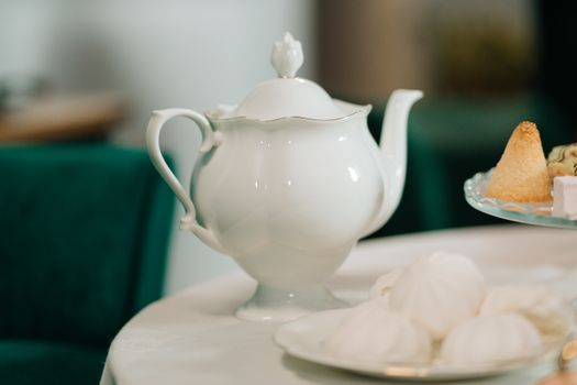 Close-up of a white porcelain teapot with assorted pastries on a table in a cozy setting.