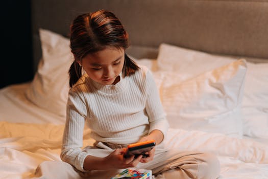 A young girl sitting cross-legged on a bed, using a smartphone in a warm indoor setting.