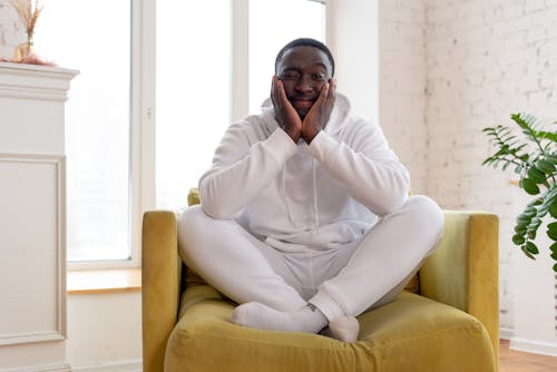 Tranquil cheerful African American male in white hoodie and trousers smiling and leaning head on hands while looking at camera