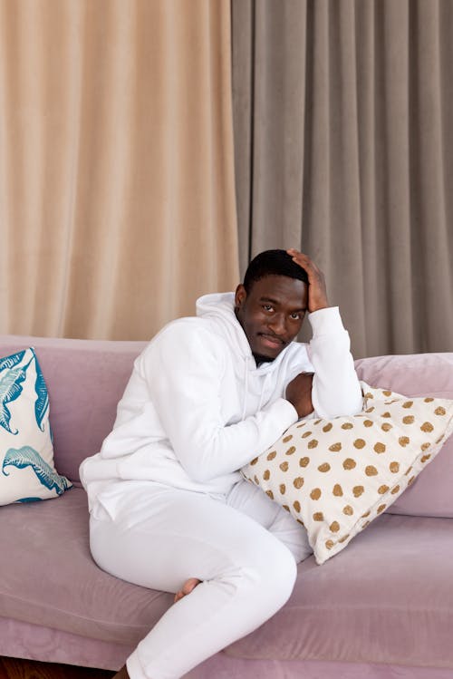 Free Cheerful African American male with short hair in white clothes sitting on comfortable pink sofa and looking at camera in living room Stock Photo