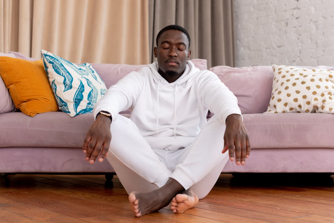 Free Calm African American male in white clothes sitting with closed eyes and crossed legs on floor against comfortable sofa in living room Stock Photo