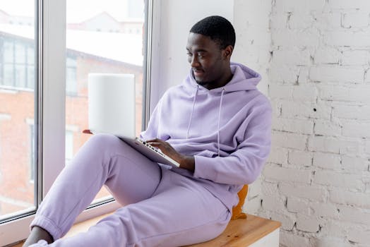 A young black man in casual wear using a laptop on a windowsill, working from home.