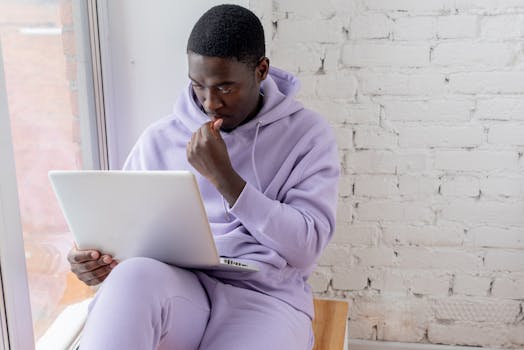 High angle of concentrated African American male remote worker sitting on windowsill and watching netbook while touching face