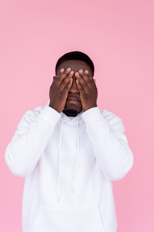 Unhappy African American male in casual sweatshirt covering face in studio against pink background