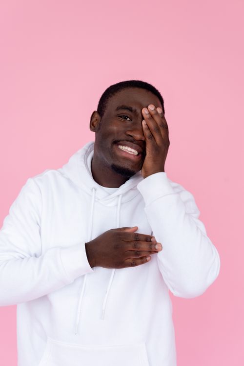 Free Cheerful African American male model wearing white hoodie covering face with hand and looking at camera against pink background Stock Photo