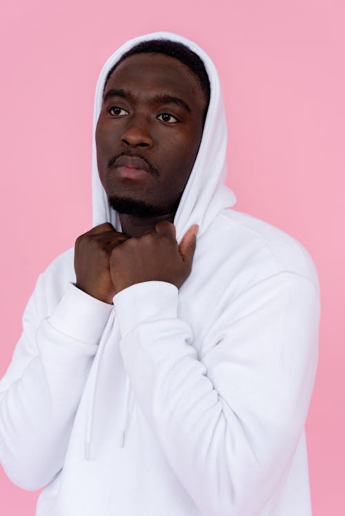 Serious African American male model in white sweatshirt keeping hands on hood and thoughtfully looking away against pink background