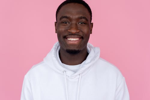 Positive African American male wearing white sweatshirt smiling widely against pink background and looking at camera