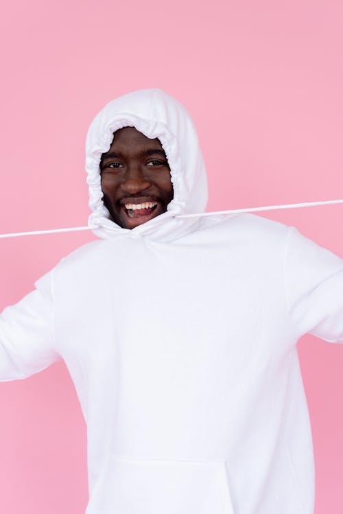 Delighted African American male in hoodie smiling happily with mouth opened against pink background and looking at camera