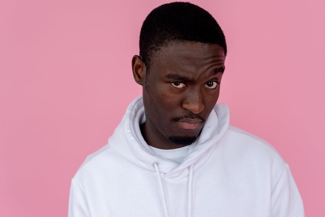 Serious African American male model wearing white sweatshirt looking at camera with unsure gaze against pink background