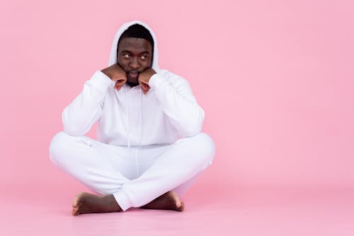 Full body of African American man with unhappy childish face sitting in studio with crossed legs against pink background