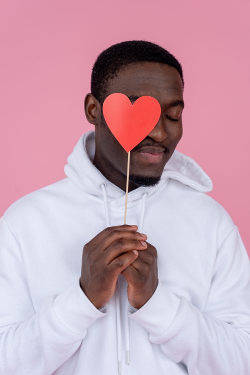 Free Young African American male in white hoodie showing red heart on stick on pink background of studio Stock Photo