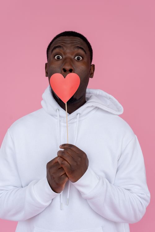 Free Surprised young African American guy in white hoodie covering mouth with pink paper heart on stick while looking at camera on pink background in light studio Stock Photo
