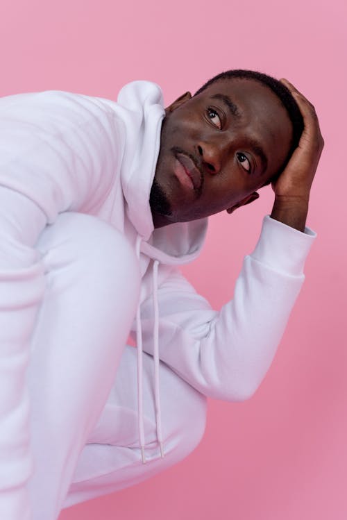Free Confident young black guy in white hoodie and pants with hand on head looking away on pink background in bright studio Stock Photo