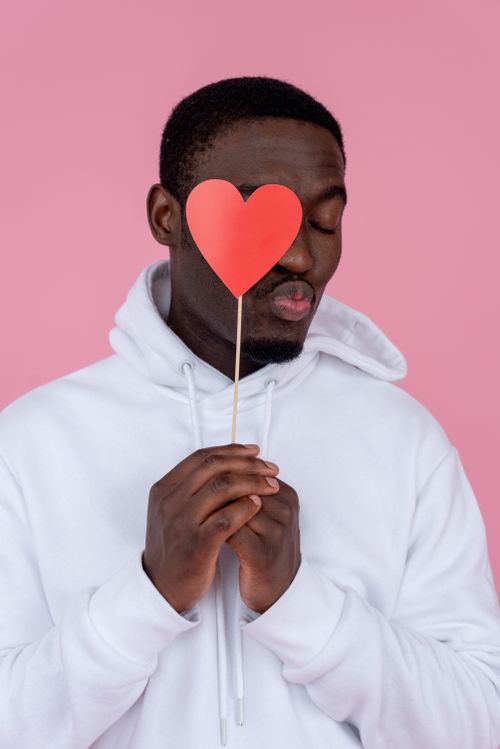 Free Young African American guy in white hoodie covering eye with red paper heart on stick while doing air kiss gesture with lips with closed eyes on pink background in studio Stock Photo
