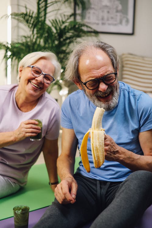 A Man Holding a Peeled Banana and a Woman Holding a Drink Sitting on Yoga Mats