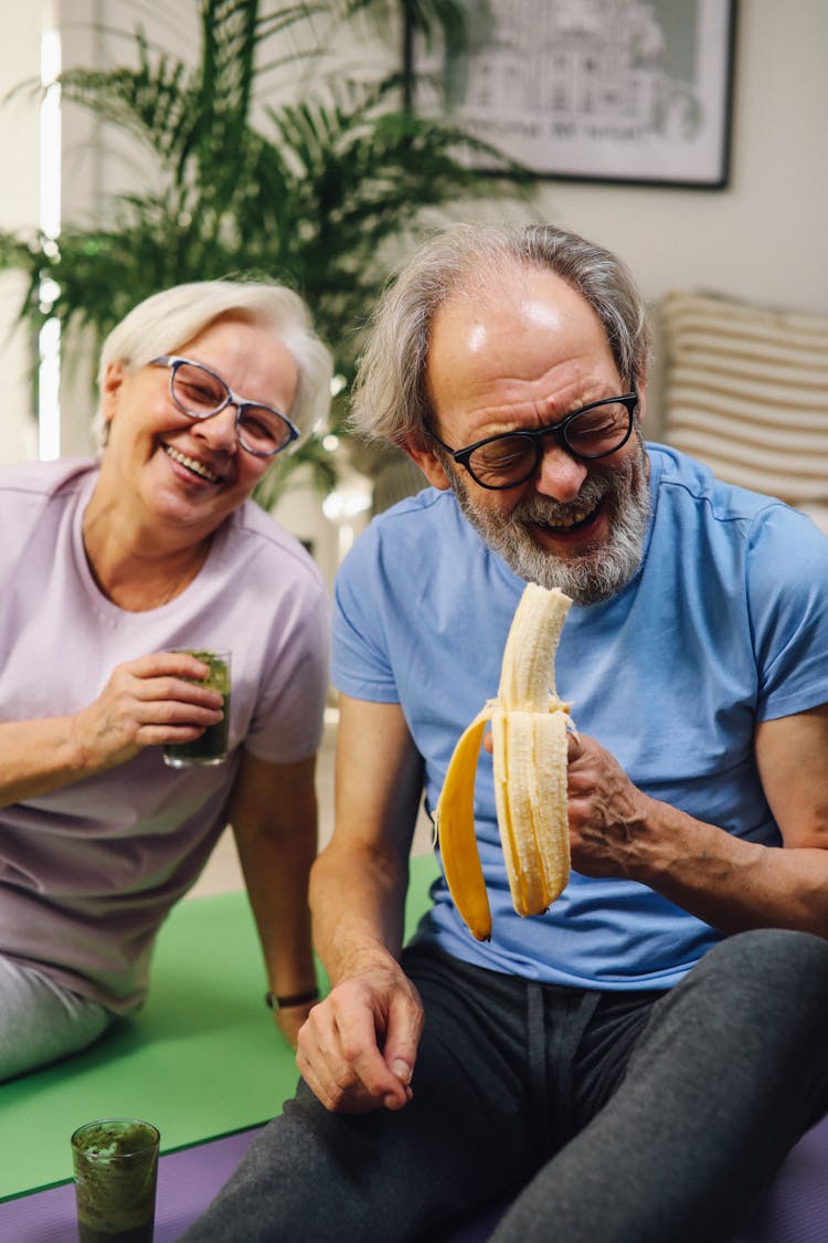 A Man Holding A Peeled Banana And A Woman Holding A Drink Sitting On Yoga Mats