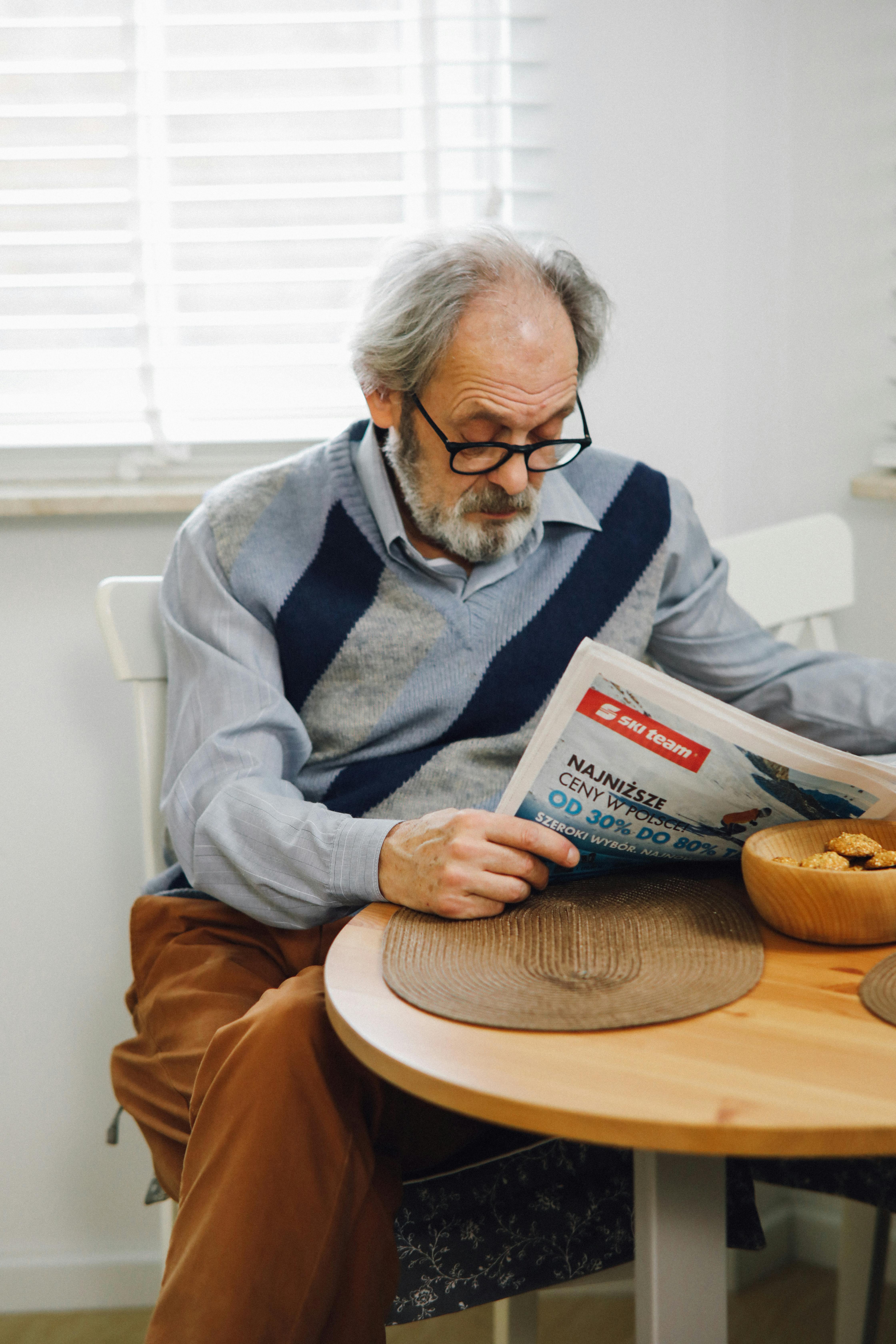 an elderly man sitting near the wooden table while reading newspaper