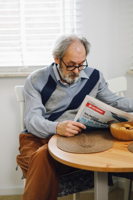 Elderly man with gray hair and glasses reading a newspaper at a wooden table indoors.
