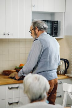 Elderly man with gray hair wearing eyeglasses, cooking in a spacious kitchen. Focus on domestic life.