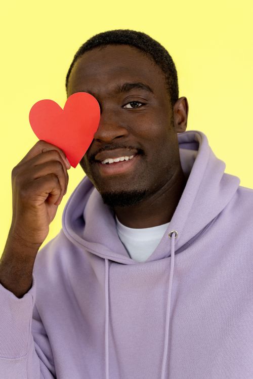 Free Cheerful man in a hoodie holding a red heart shape against a vibrant yellow backdrop. Stock Photo