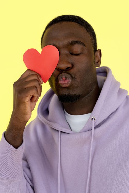 Free A man poses playfully with a heart cutout against a bright yellow background, wearing a casual hoodie. Stock Photo