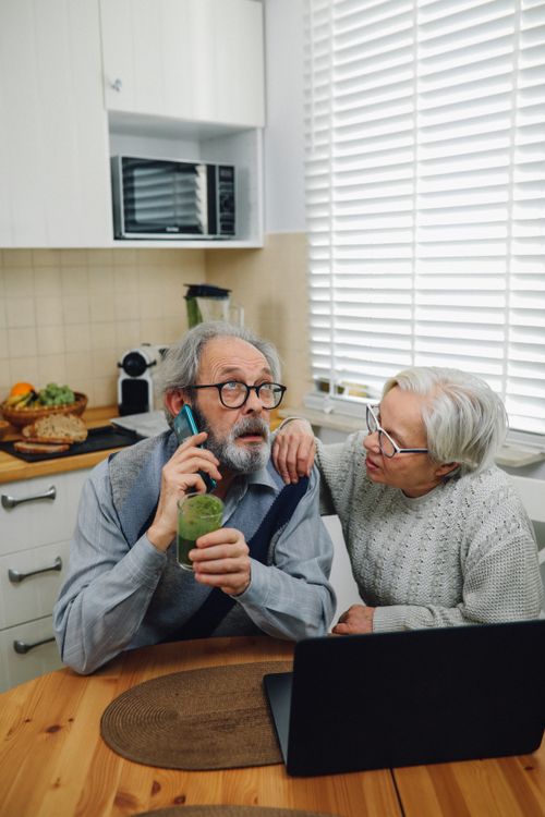 Free Senior couple in a cozy kitchen, discussing while using a phone and laptop. Stock Photo