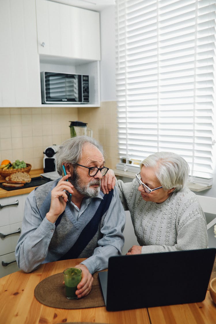 Elderly Couple Sitting By A Table Using A Phone And A Laptop