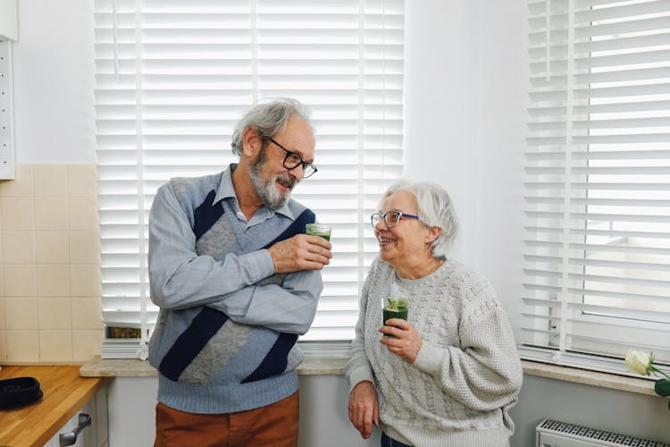 Happy Senior Couple Standing In Kitchen With Smoothie Drinks 