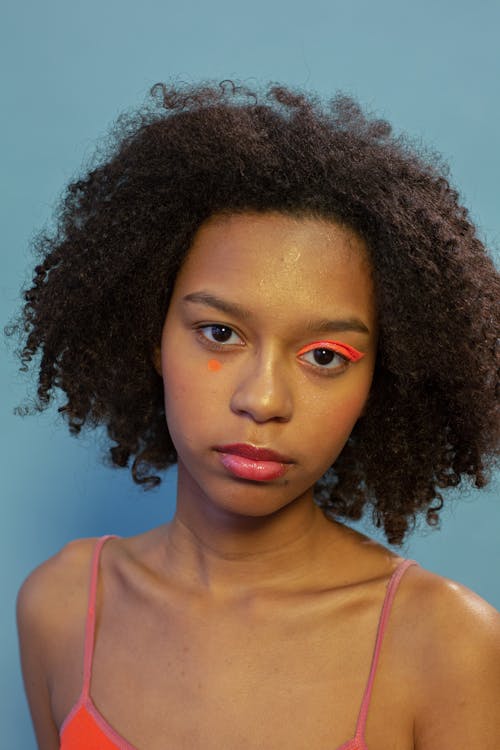 Young calm African American female model with curly hair and shiny orange eyeshadow on one eye looking at camera against blue wall