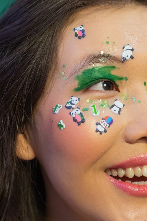Crop joyful Asian female model with creative vivid makeup and panda stickers on flawless face looking away in studio