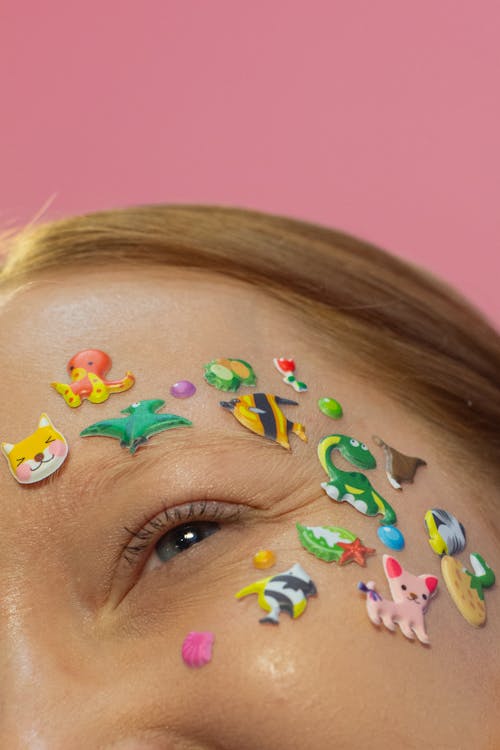 Crop glad redhead female model with colorful small stickers in shape of animals on face looking away on pink background in studio